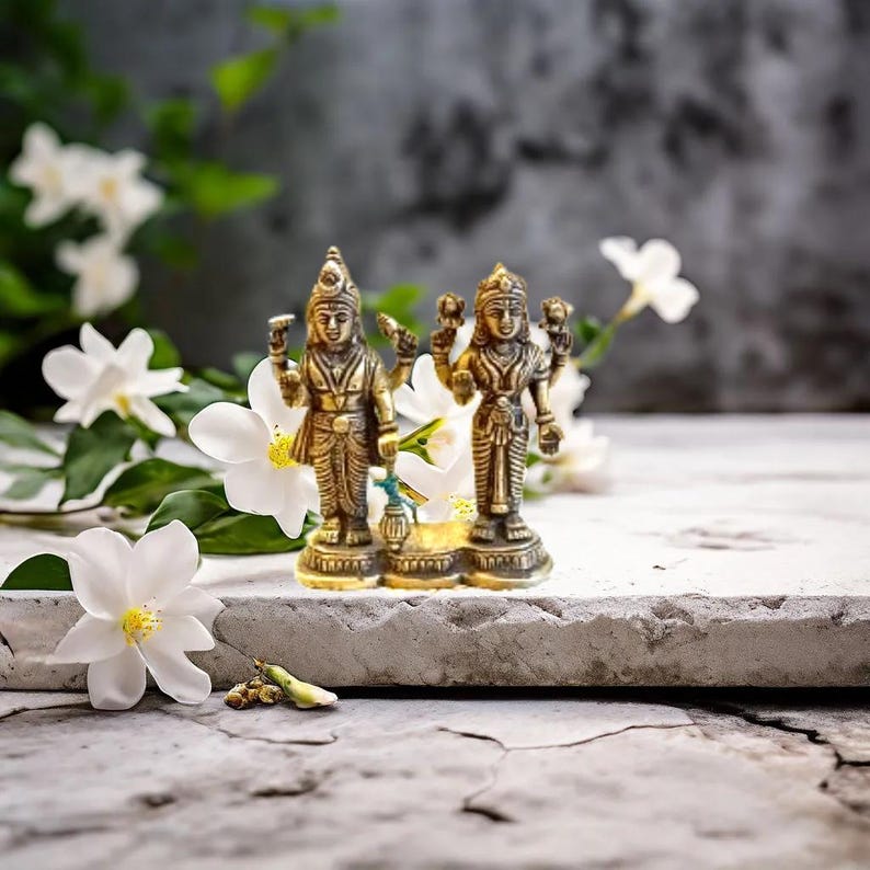 May include: A gold-colored statue of a Hindu deity couple, standing on a white surface with white flowers in the background.