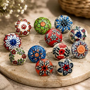 May include: A collection of eleven round ceramic cabinet knobs featuring various hand-painted floral and geometric patterns in vibrant colours including red, blue, green, orange, and black on white or coloured backgrounds with metallic hardware.