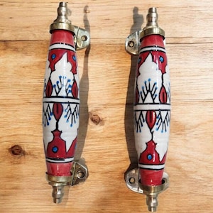 May include: Two decorative cabinet handles with a red, white, and blue floral design. The handles are cylindrical with brass hardware and are mounted on a wooden surface. The handles are approximately 6 inches long.