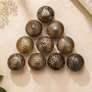 Op de afbeelding: Een verzameling van tien decoratieve kastknoppen met ingewikkelde gouden en zwarte patronen. De knoppen zijn gerangschikt in een driehoek op een crèmekleurig oppervlak, met bloemaccenten op de achtergrond. Elke knop heeft een uniek ontwerp.
