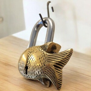 May include: A decorative padlock with a fish-shaped brass body and a silver-colored shackle. The fish has detailed scales and fins, and a keyhole is visible on the side. The lock is resting on a wooden surface.