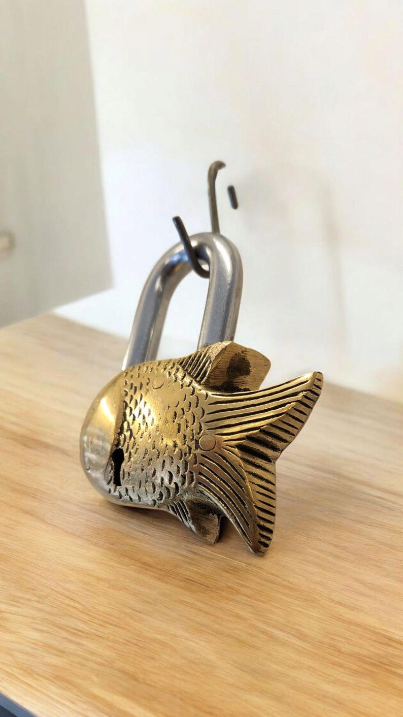 May include: A decorative padlock with a fish-shaped brass body and a silver-colored shackle. The fish has detailed scales and fins, and a keyhole is visible on the side. The lock is resting on a wooden surface.