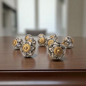 May include: A collection of decorative ceramic knobs with a floral design. The knobs are primarily white with black and gray patterns, accented with gold-colored centers. They are arranged on a dark brown wooden surface.