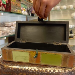 Puede incluir: Una caja de madera con tapa con bisagras y un dise&ntilde;o geom&eacute;trico colorido. La caja est&aacute; abierta y el interior est&aacute; vac&iacute;o.