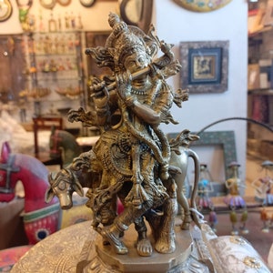 May include: A gold-colored metal statue of a Hindu deity playing a flute, standing on a decorative base. The deity is depicted with multiple arms and is riding a cow.