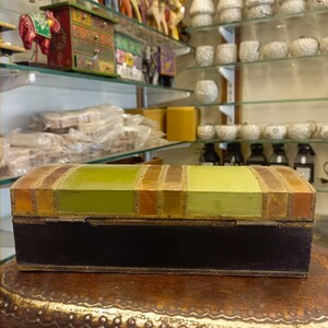 Puede incluir: Una peque&ntilde;a caja rectangular de madera con una base negra y una tapa rayada en verde y dorado. La caja tiene una tapa con bisagras y est&aacute; decorada con detalles dorados.