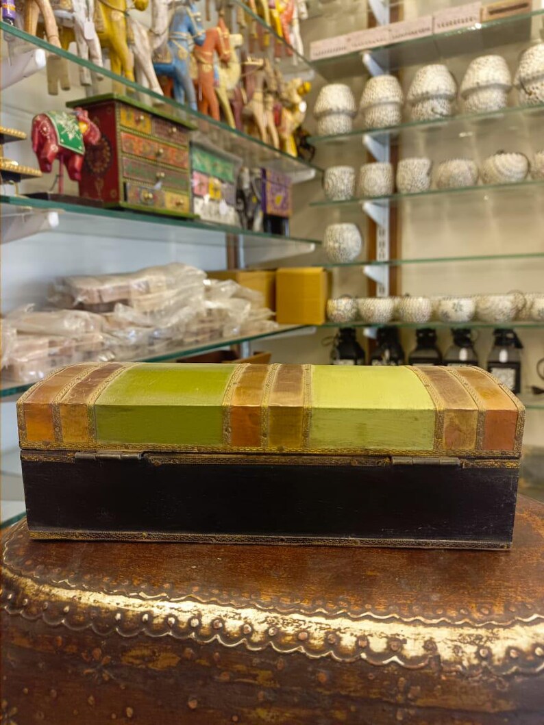 Puede incluir: Una peque&ntilde;a caja rectangular de madera con una base negra y una tapa rayada en verde y dorado. La caja tiene una tapa con bisagras y est&aacute; decorada con detalles dorados.