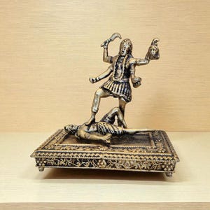 May include: A bronze-coloured statue of a multi-armed deity standing on a figure, holding a sword and a severed head. The statue is on a decorative, rectangular base with ornate detailing.