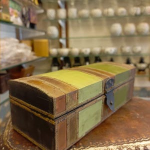 Puede incluir: Una caja de madera de estilo vintage con tapa con bisagras y cierre de metal. La caja est&aacute; pintada en tonos de verde, marr&oacute;n y negro, con un patr&oacute;n a rayas. La caja est&aacute; sentada sobre una superficie de cuero marr&oacute;n.