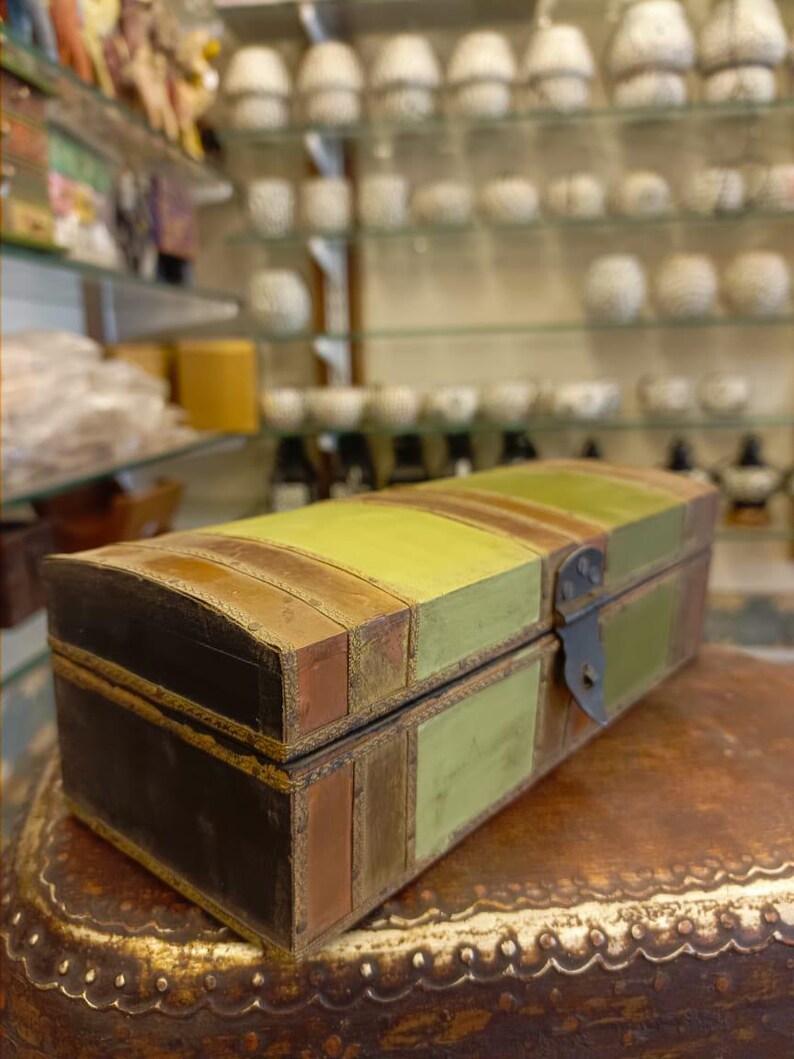 Puede incluir: Una caja de madera de estilo vintage con tapa con bisagras y cierre de metal. La caja est&aacute; pintada en tonos de verde, marr&oacute;n y negro, con un patr&oacute;n a rayas. La caja est&aacute; sentada sobre una superficie de cuero marr&oacute;n.