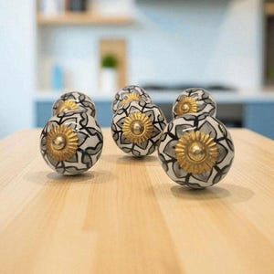 Puede incluir: Un conjunto de tiradores decorativos para armarios con un diseño floral. Los tiradores son blancos con un patrón negro y gris y un centro dorado. Están dispuestos sobre una superficie de madera clara, con un fondo de cocina borroso.