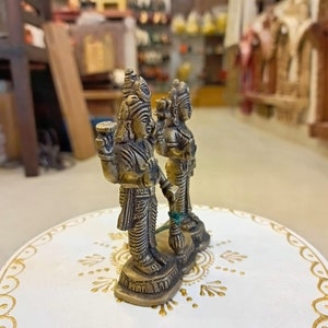 May include: Two small gold-colored statues of deities standing on a white circular base with gold accents. The statues are intricately detailed and have a traditional Indian style.