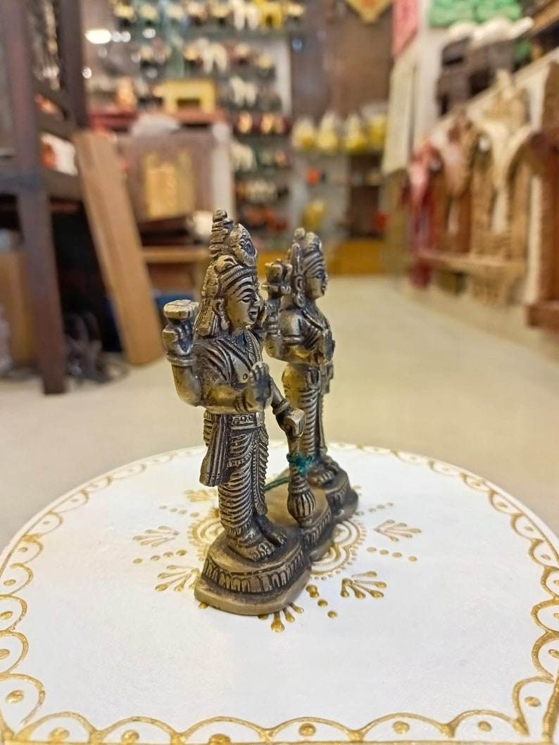 May include: Two small gold-colored statues of deities standing on a white circular base with gold accents. The statues are intricately detailed and have a traditional Indian style.
