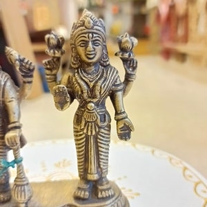 May include: A gold-colored metal statue of a Hindu deity with four arms, holding a lotus flower in each hand. The statue is standing on a base with intricate details.