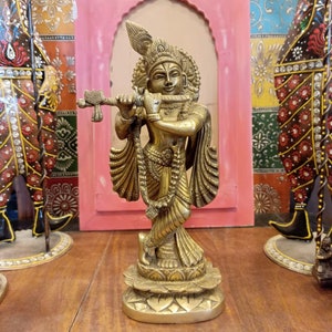 May include: A gold-colored statue of Krishna playing a flute. The statue is standing on a lotus-shaped base and is decorated with intricate details. The statue is made of brass and is a traditional Hindu religious artifact.