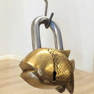 May include: A decorative brass padlock shaped like a fish. The fish-shaped body has detailed scale patterns and a keyhole. The shackle is made of silver-colored metal. The padlock is hanging from a hook.