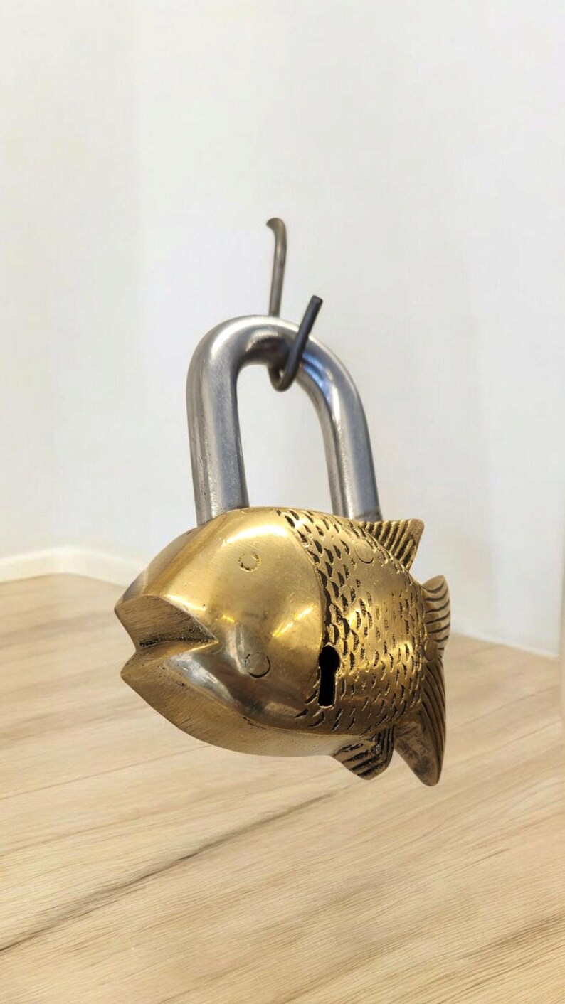 May include: A decorative brass padlock shaped like a fish. The fish-shaped body has detailed scale patterns and a keyhole. The shackle is made of silver-colored metal. The padlock is hanging from a hook.