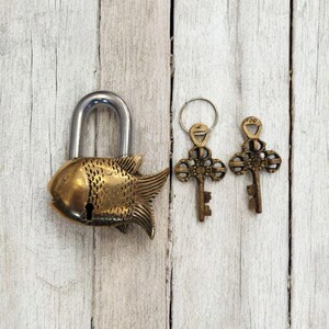 May include: A brass fish-shaped padlock with a silver shackle, accompanied by two ornate keys on a ring. The keys and lock are detailed with decorative patterns. The items rest on a weathered, light-colored wooden surface.