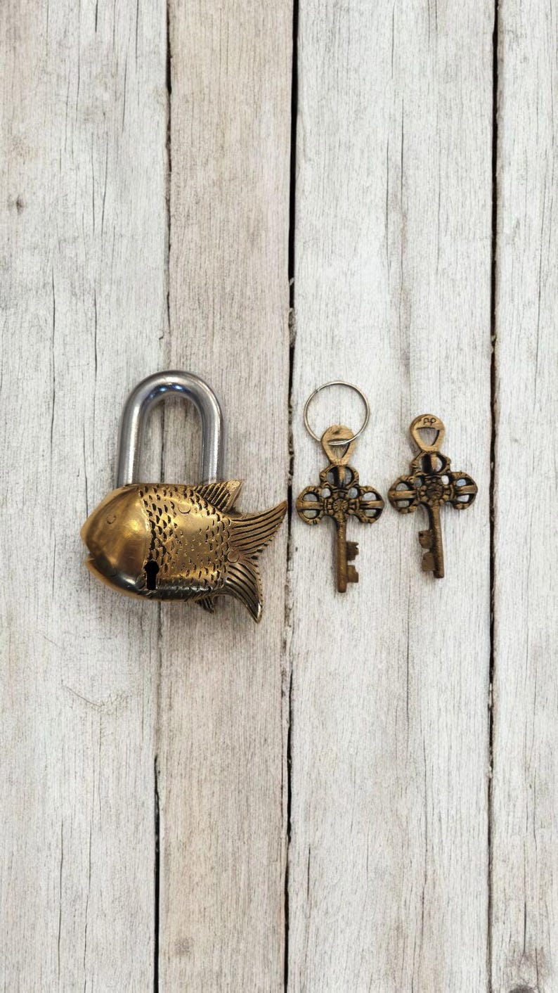 May include: A brass fish-shaped padlock with a silver shackle, accompanied by two ornate keys on a ring. The keys and lock are detailed with decorative patterns. The items rest on a weathered, light-colored wooden surface.