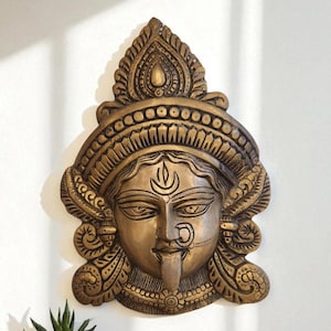 Puede incluir: Una escultura mural de color bronce que representa el rostro de una deidad con detalles intrincados. La escultura presenta una expresión serena, un tocado ornamentado y piercings faciales. Una pequeña planta suculenta en maceta se encuentra sobre una superficie de madera.