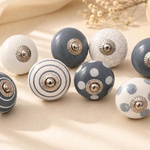 May include: A collection of eight round ceramic cabinet knobs featuring various designs including solid white, solid dark gray, white with gray stripes, gray with white polka dots, white with gray polka dots, and a white knob with a gray crackle finish.
