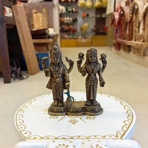 May include: A pair of small, gold-colored metal statues of Hindu deities, Shiva and Parvati, standing on a white pedestal with gold accents.
