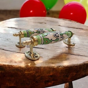 Puede incluir: Dos tiradores decorativos para armarios con un diseño floral blanco y verde. Los tiradores son de cerámica y latón, y se exhiben sobre una mesa de madera rústica. Globos rojos, verdes y amarillos en el fondo.