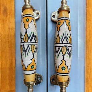May include: Two decorative cabinet handles. The handles are cylindrical with a cream base, decorated with yellow and green geometric patterns. They have brass-coloured metal accents and are mounted on a light blue cabinet door.