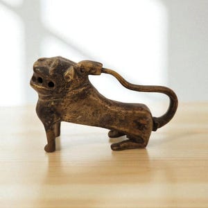 Puede incluir: Un abridor de botellas con forma de león de color bronce. El abridor está en posición agachada con un asa curva que se extiende desde su espalda. El león tiene detalles y está hecho de metal. El abridor está sobre una superficie de madera.