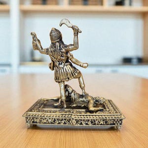 May include: A detailed brass figurine of a multi-armed deity standing over a figure on a decorative rectangular base. The deity is holding weapons and a small object. The figurine is set on a wooden table.