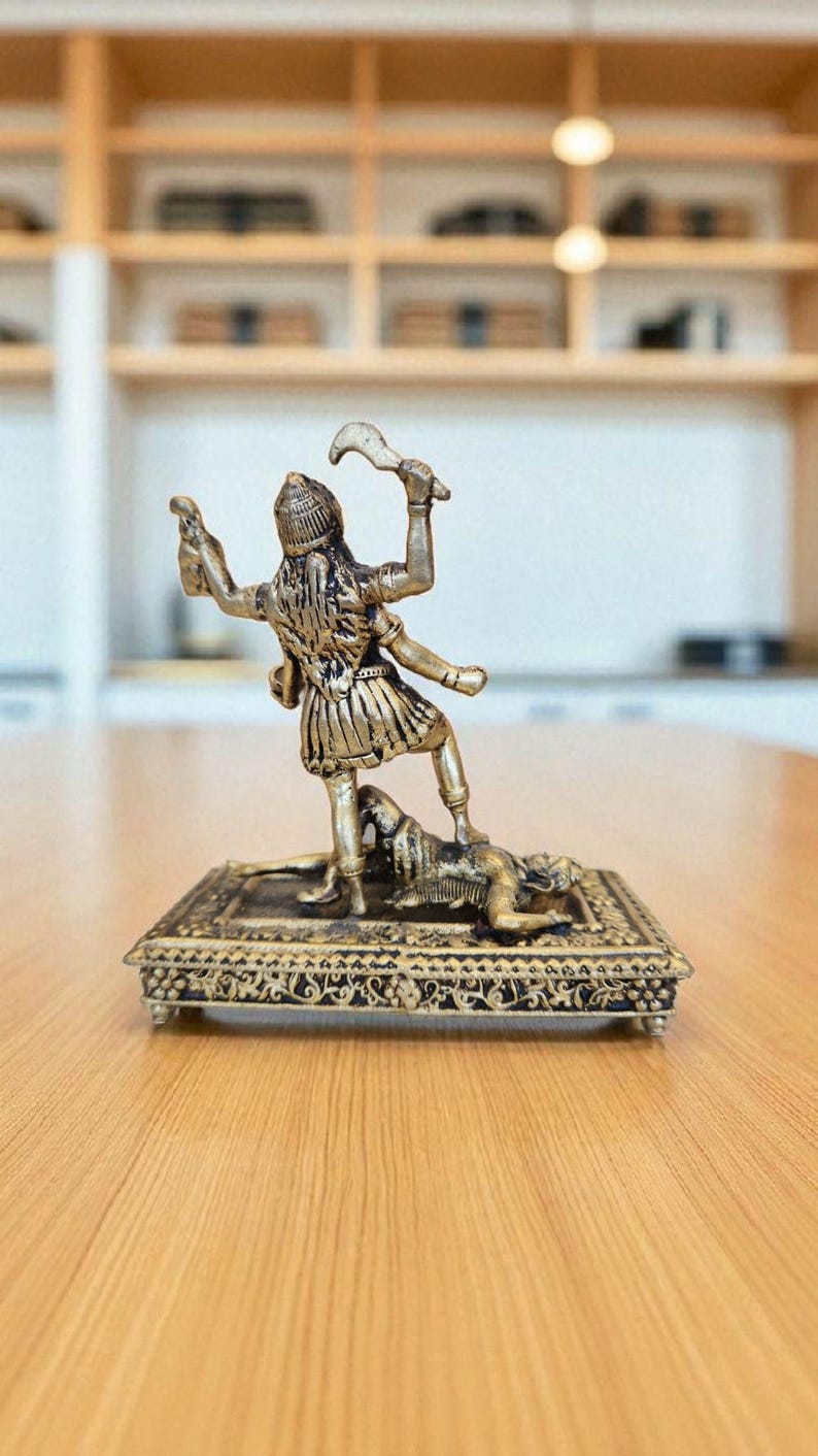 May include: A detailed brass figurine of a multi-armed deity standing over a figure on a decorative rectangular base. The deity is holding weapons and a small object. The figurine is set on a wooden table.