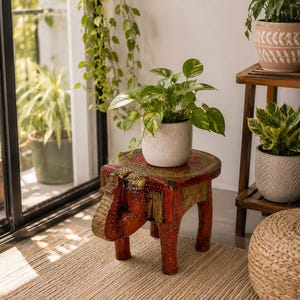 May include: A red and gold hand-carved wooden elephant stool with an oval top, featuring intricate embossed patterns and a trunk that forms one of the legs. A white speckled ceramic pot containing a green pothos plant sits on the stool.