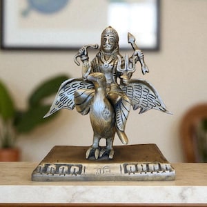 May include: A detailed silver-toned statue of a deity riding a bird, possibly Garuda. The statue is on a rectangular base with etched details. The background includes a wooden cabinet and artwork.