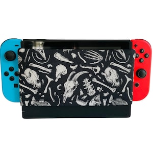 Puede incluir: Una funda de tela negra y blanca para una consola Nintendo Switch. La funda presenta un diseño de esqueletos blancos sobre fondo negro. La funda está unida a una consola Nintendo Switch azul y roja.
