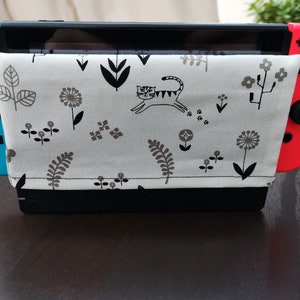 May include: A white and gray patterned fabric case with a black cat design, fits a Nintendo Switch console, and is attached to a black stand.