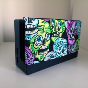 May include: A black Nintendo Switch console with a colourful skull-patterned case. The case features green, purple, blue, and black skulls with white eyes.