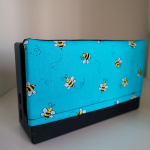 Puede incluir: Una funda azul para Nintendo Switch con una base negra. La funda está cubierta de una tela con un patrón repetitivo de abejas amarillas y negras sobre un fondo turquesa.
