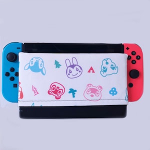 Könnte beinhalten: Eine weiße Nintendo Switch Konsolenhaut mit einem bunten Animal Crossing Muster. Die Haut zeigt verschiedene Charaktere und Symbole aus dem Spiel, darunter einen Palmenbaum, ein Zelt und eine Glocke. Die Haut ist an einer schwarzen Nintendo Switch Konsole befestigt.