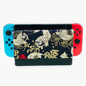 Puede incluir: Una consola Nintendo Switch azul y roja con una funda negra y dorada con un patrón de peces koi. La funda tiene una solapa que cubre la pantalla.