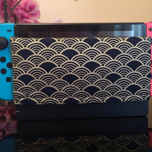 Pode incluir: Um console Nintendo Switch azul e vermelho com uma capa de padrão preto e dourado. A capa tem um padrão repetitivo de semicírculos sobrepostos.