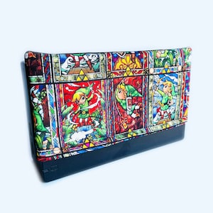 Puede incluir: Una funda negra y blanca para Nintendo Switch con un diseño de vitral que presenta personajes de la serie de videojuegos The Legend of Zelda. La funda tiene un diseño colorido con acentos rojos, amarillos, verdes, azules y morados.