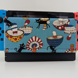 May include: A blue and black Nintendo Switch console with a custom case featuring a whimsical design of black cats, mushrooms, and flowers. The case is made of fabric and has a blue background with white and red accents.