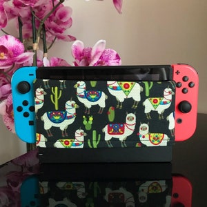 May include: A black Nintendo Switch console with a colorful llama-themed case. The case features white llamas with colorful geometric patterns and green cacti on a black background.