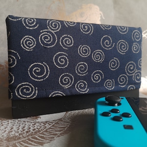 Puede incluir: Una funda de tela azul marino con un diseño de espirales color crema. La funda es rectangular y se apoya sobre una base negra. Un mando de juego turquesa está en primer plano.
