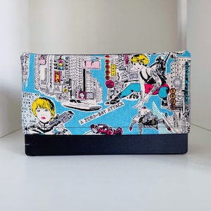 Puede incluir: Un embrague de tela azul y blanco con una ciudad y personajes de dibujos animados. El texto "A Zero-Day Attack" está impreso en la parte delantera.