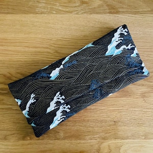 Pode incluir: Um item retangular de tecido dobrado com fundo preto e um padrão de ondas repetido em branco e azul claro. As ondas são estilizadas, com linhas douradas adicionando detalhes. O item está sobre uma superfície de madeira.