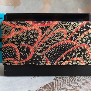 May include: A blue Nintendo Switch console with a black and red patterned case. The case has a gold design and is made of fabric.