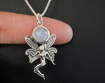 May include: A silver necklace featuring a fairy pendant with a moonstone. The fairy has detailed wings and a delicate form. The moonstone is round and set above the fairy's head. The chain is a fine, square link style.