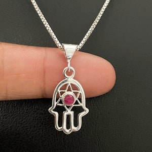 May include: A silver chain necklace with a hamsa hand pendant featuring a red gemstone in the centre. The hamsa hand is a popular symbol of protection and good luck.