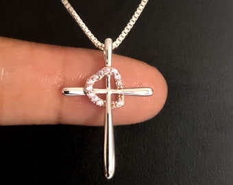 Puede incluir: Un collar de cadena de plata con un colgante de cruz de plata. La cruz tiene forma de corazón con piedras de circonita cúbica rosas.